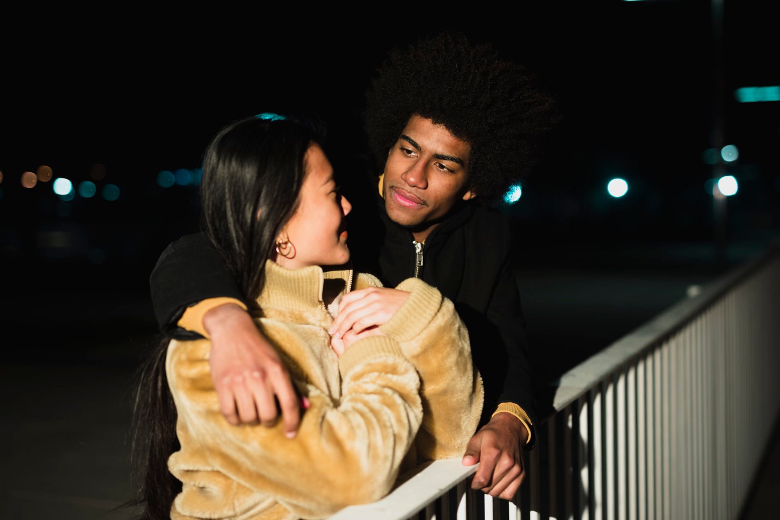 A couple stands close together at night, with warm lights twinkling in the background, showcasing a loving moment.