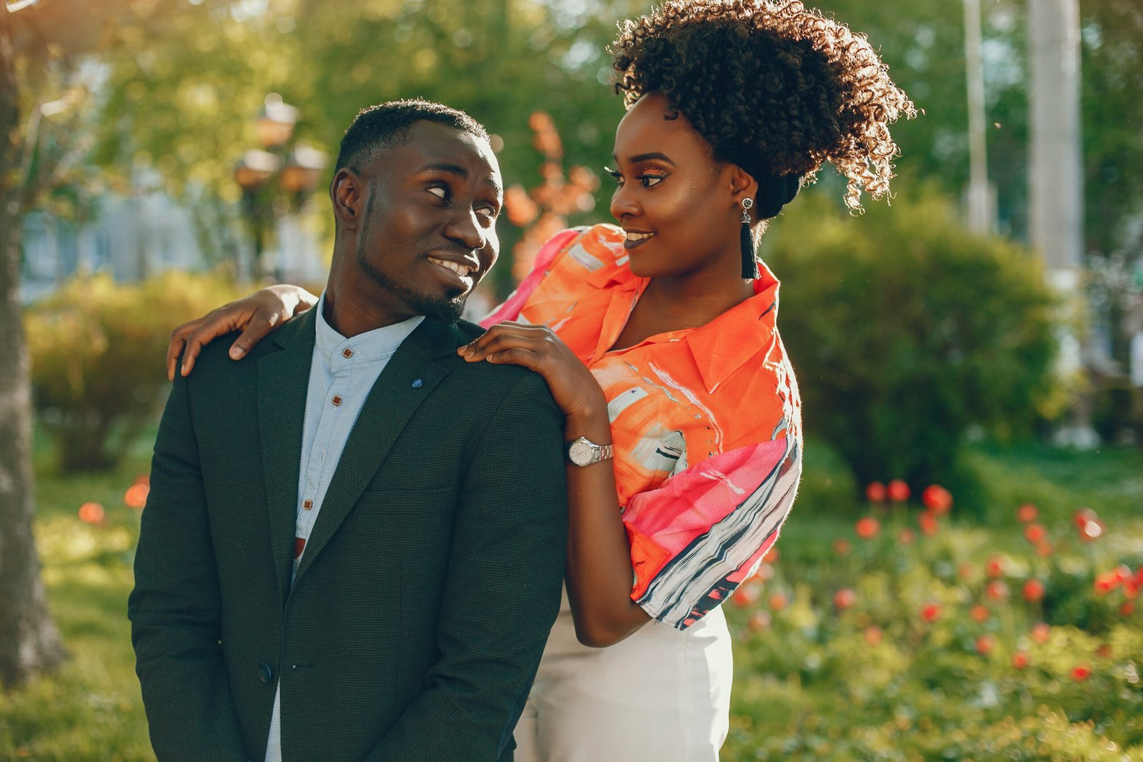 A couple stands close together in a sunlit park, surrounded by greenery and blooming flowers, with one person playfully resting a hand on the other's shoulder.